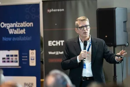 A speaker addressing the room with a handheld microphone, standing between the partner roll-up banners.